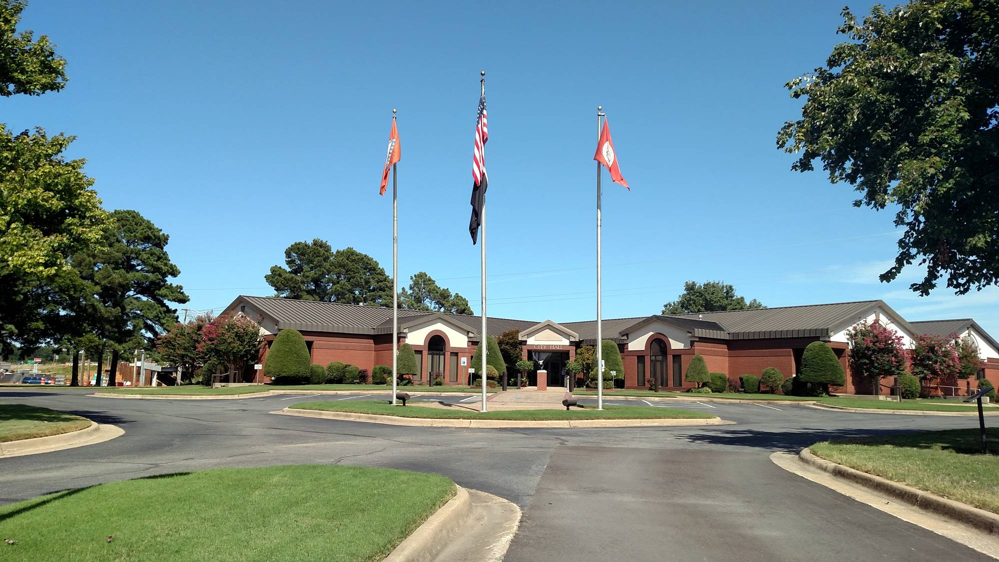 A large brick building with a dark roof, surrounded by neatly trimmed trees and grass, stands behind three flagpoles flying the U.S. flag and two red flags on a clear, sunny day. A circular driveway is in front.