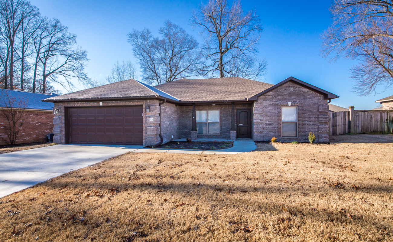 A single-story brick house with a brown shingled roof, attached two-car garage, and leafless trees in the background. The yard has brown grass, and a wooden fence is on the right. The sky is clear and blue.