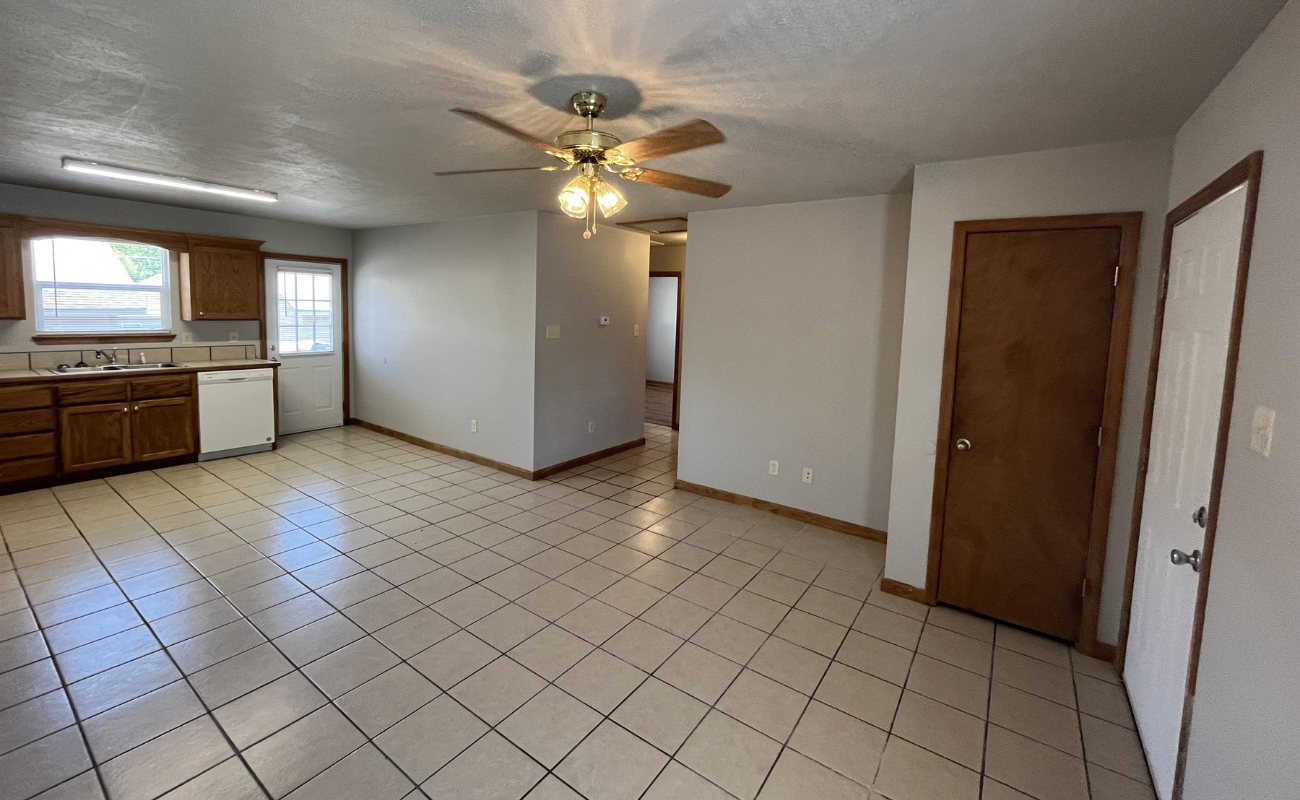 25 Marie Lane Spacious kitchen and dining area with tiled floor, wooden cabinets, a white dishwasher, and a ceiling fan with lights. There is a wooden door on the right and natural light coming through a window over the sink.