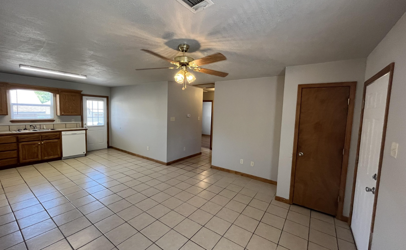 A small kitchen and dining area with tiled floors. The kitchen has wooden cabinets, a dishwasher, and a window above the sink. A ceiling fan with a light is in the center, and there are wooden doors on the right.