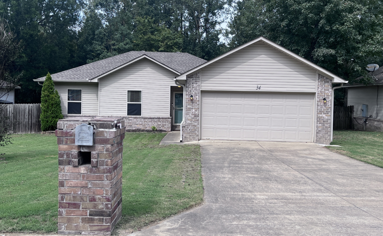 A single-story house with light-colored siding and brick accents. It features a two-car garage and a driveway. A brick mailbox stands by the entrance. The yard has neatly trimmed grass, and trees are visible in the background.