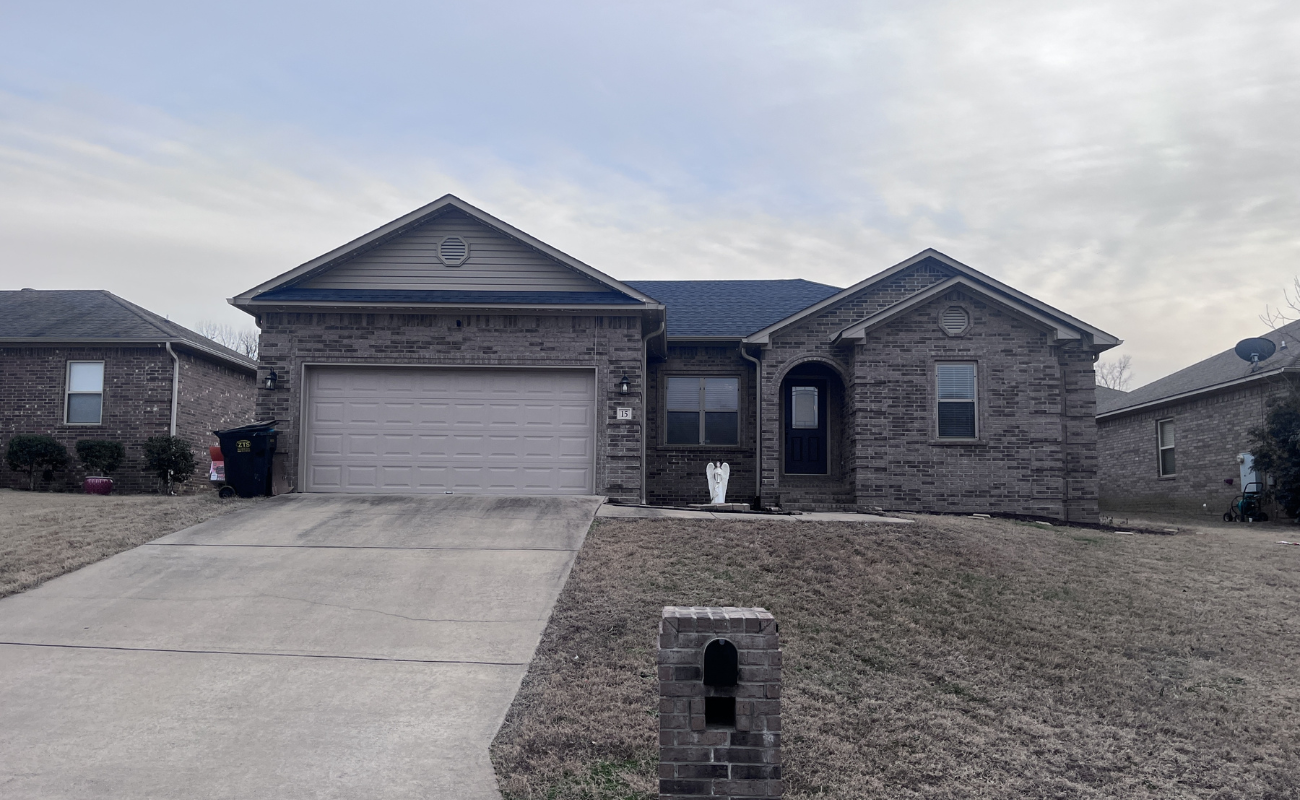 A single-story brick house with a two-car garage and arched front entryway. It has a concrete driveway and a small mailbox at the front. The lawn is slightly patchy and the sky is overcast.