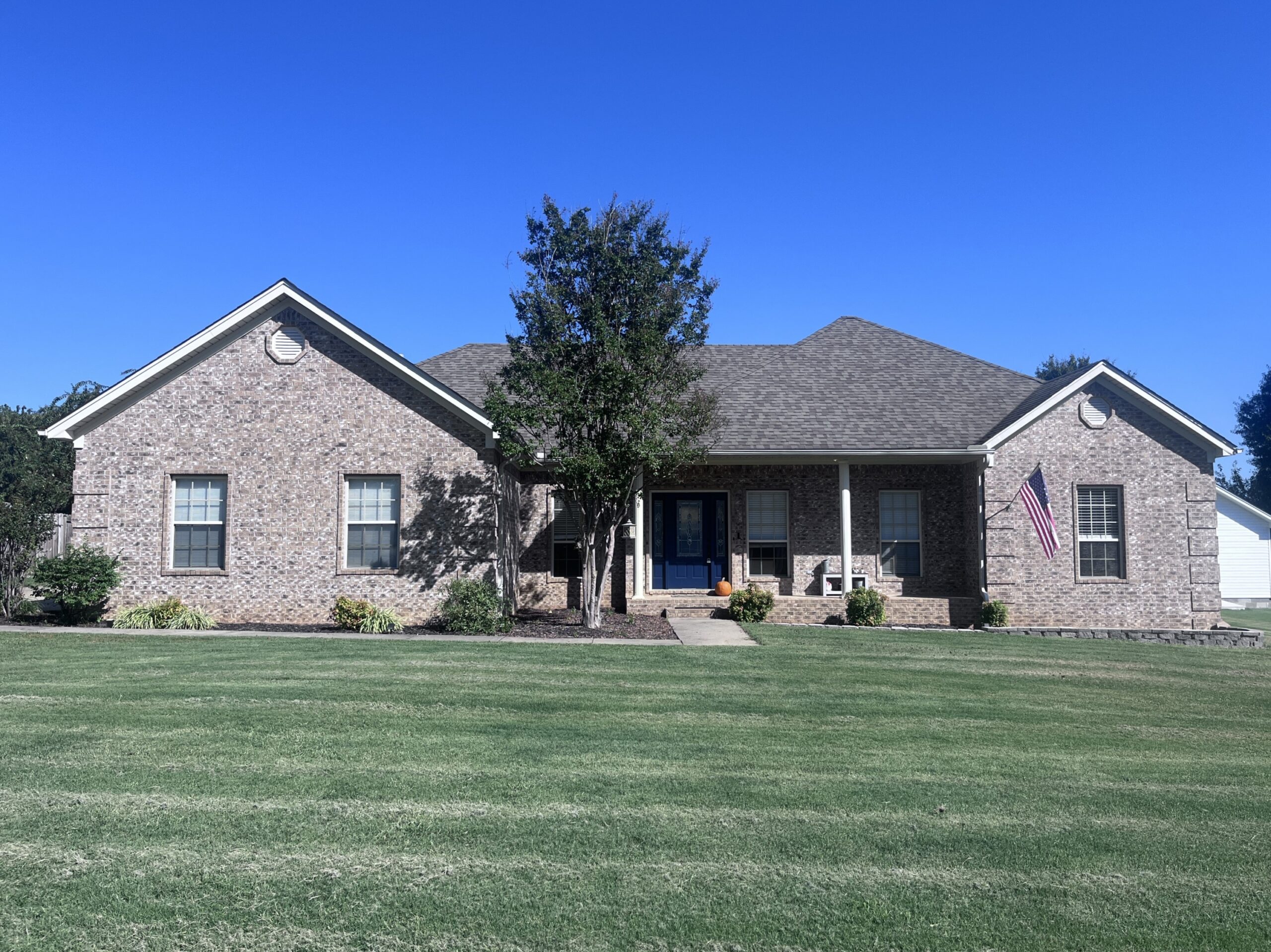 7a9d0eb0ca4d41f1b7a6e726f3516ce9 Single-story brick house with a covered front porch, blue front door, and columns. A tree is planted in front, and the lawn is neatly mowed. An American flag hangs by the porch on the right. The sky is clear and blue.