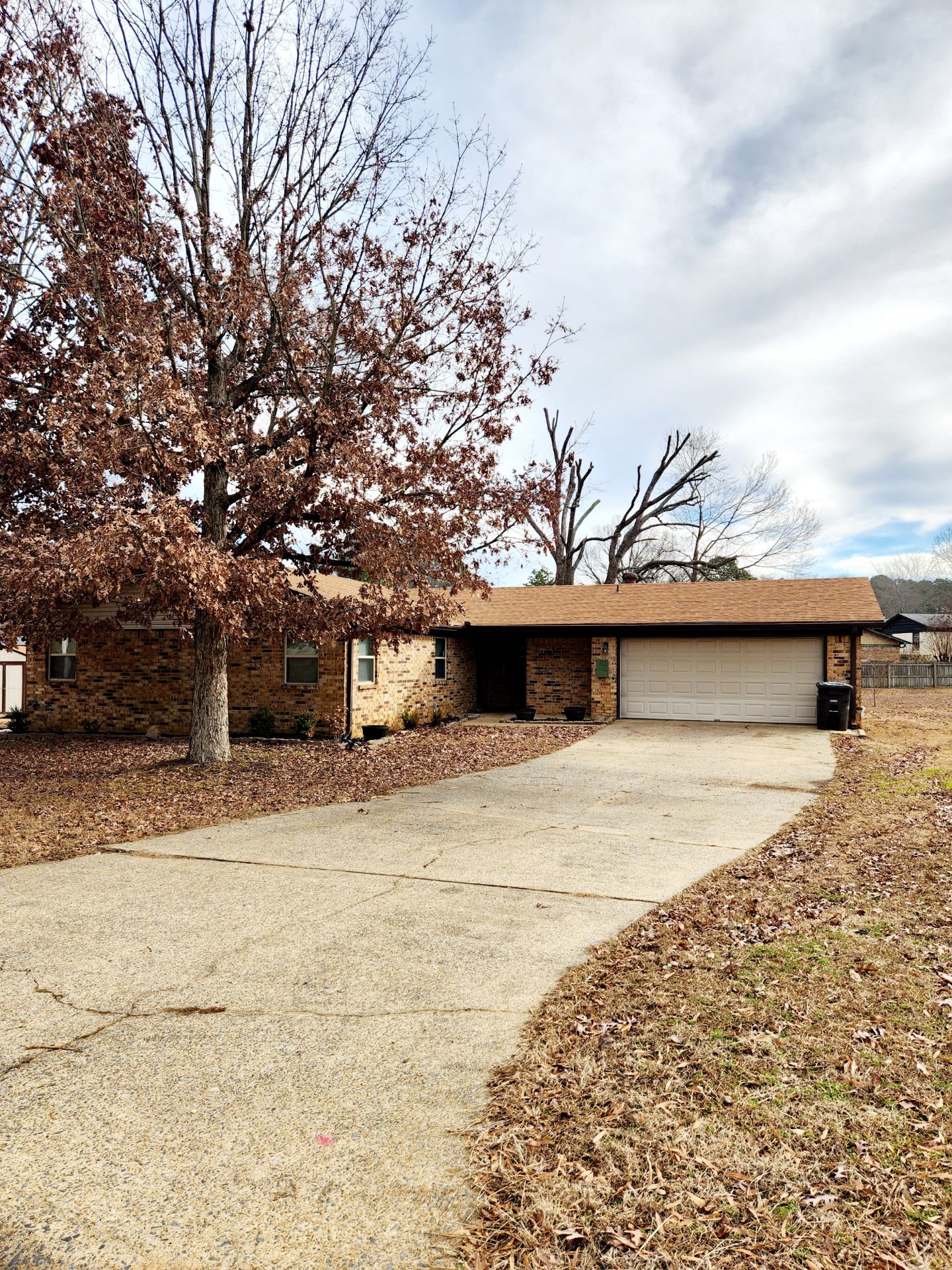 934958447539403089461d0a8cf6ee9f A single-story brick house with a two-car garage, a wide curved driveway, and large leafless trees in the yard under a cloudy sky.
