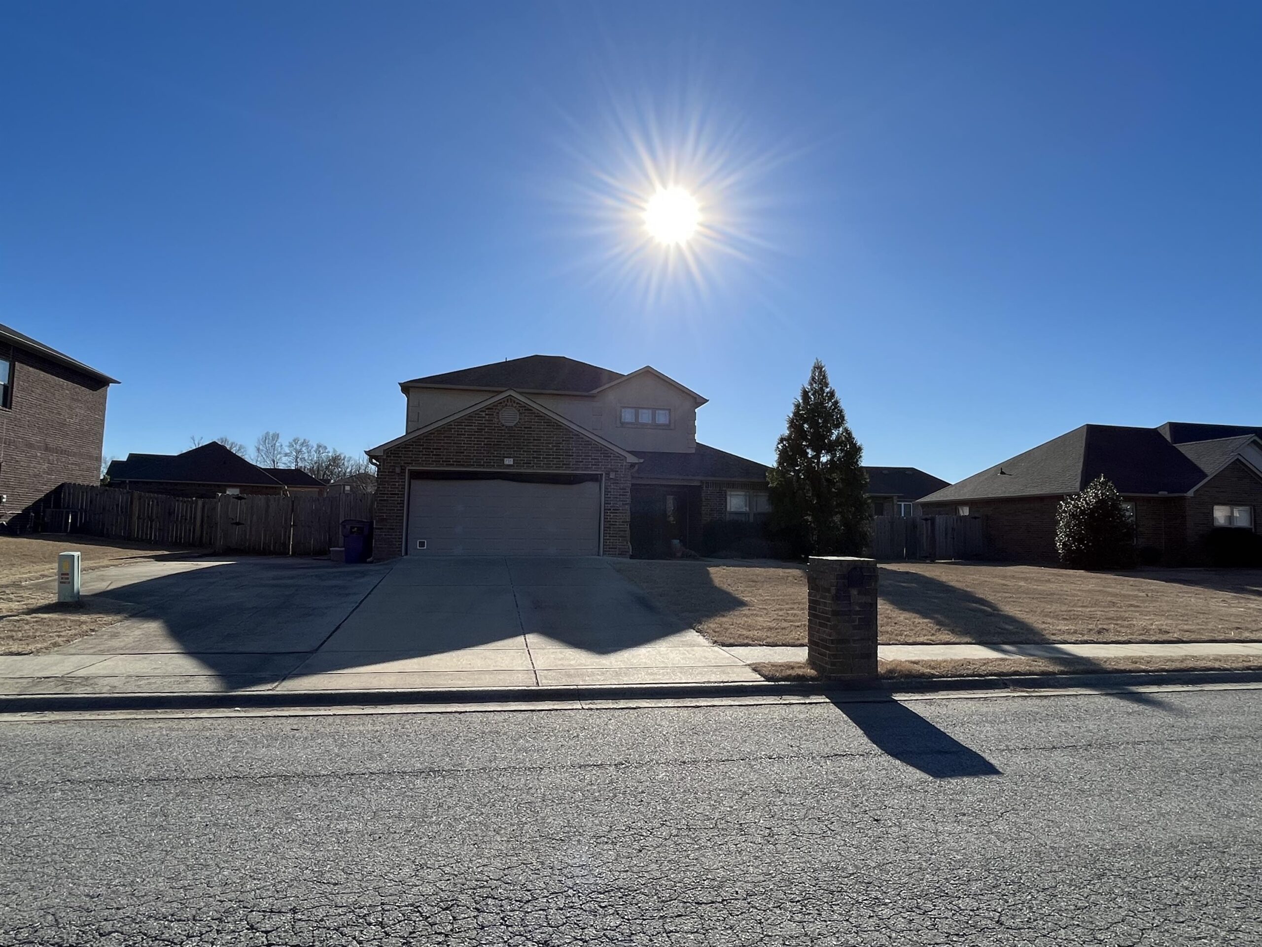 de7860e40efe4df0bc5204ac0cf36550 A two-story brick house with a driveway and garage, under a clear blue sky with the sun shining brightly above, casting long shadows on the pavement and lawn.
