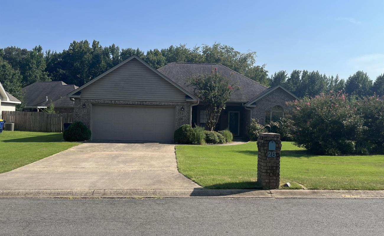 28 Hillsboro Drive Cabot A single-story suburban house with a two-car garage, gray siding, and brick accents. It has a wide driveway, a mailbox on a brick post, green lawn, shrubs, and trees in the background under a clear blue sky.