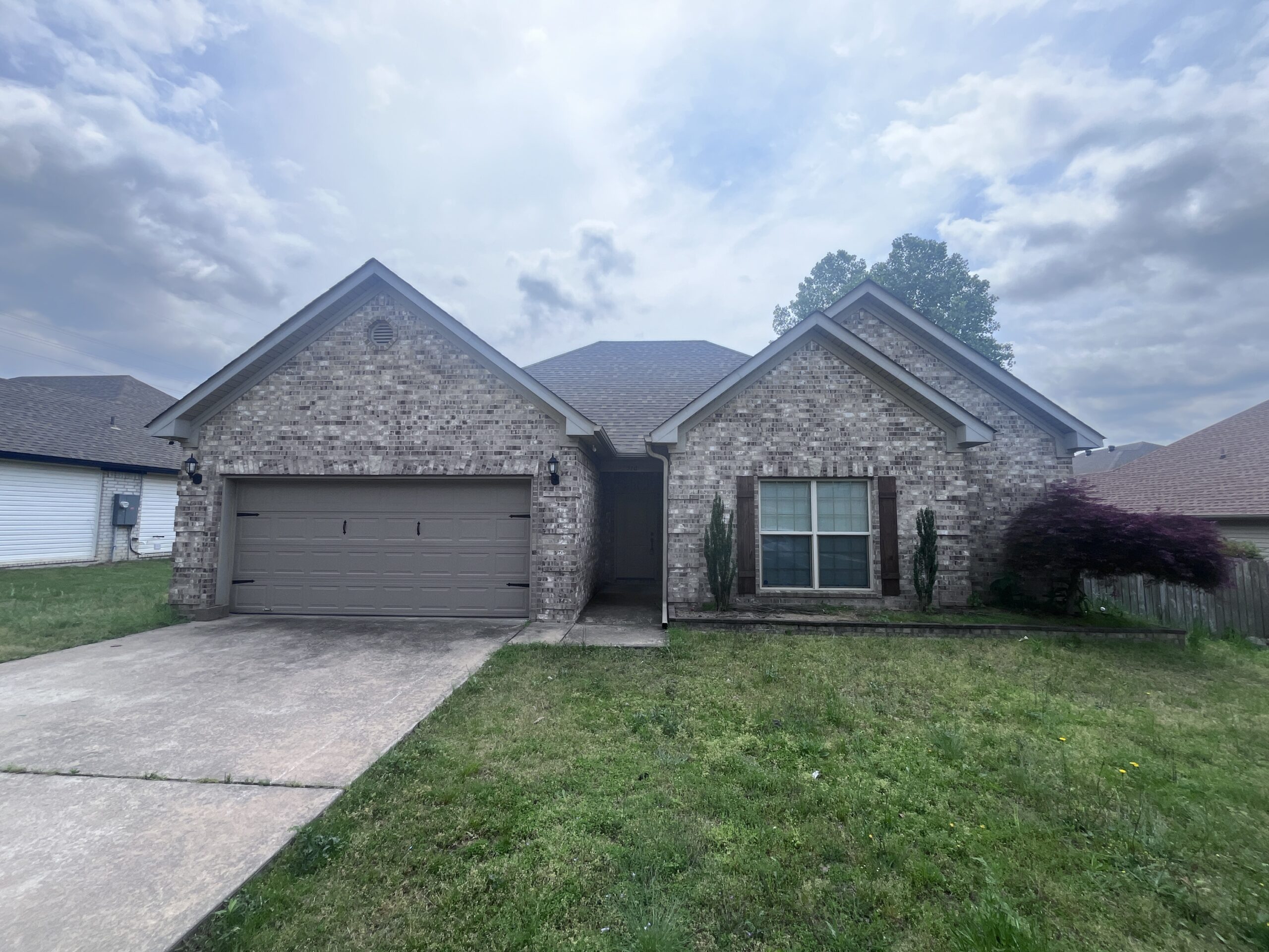 3b4f68bc341e43869990854652cecac6 A single-story brick house with a two-car garage, front lawn, and two tall windows. The sky is mostly cloudy, and there are neighboring houses on either side.
