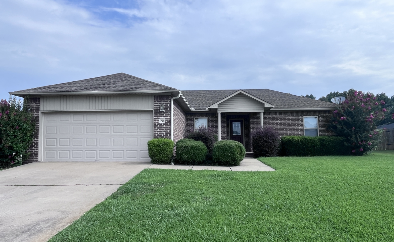 A single-story brick house with a two-car garage, neatly trimmed bushes in front, and a well-maintained green lawn under a partly cloudy sky.