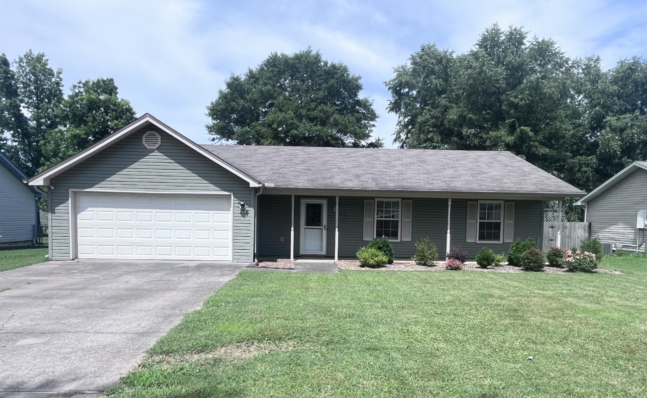 A single-story house with gray siding, a two-car garage, and a covered front porch. The house has a neatly trimmed lawn, small bushes, and a concrete driveway, with trees in the background under a partly cloudy sky.