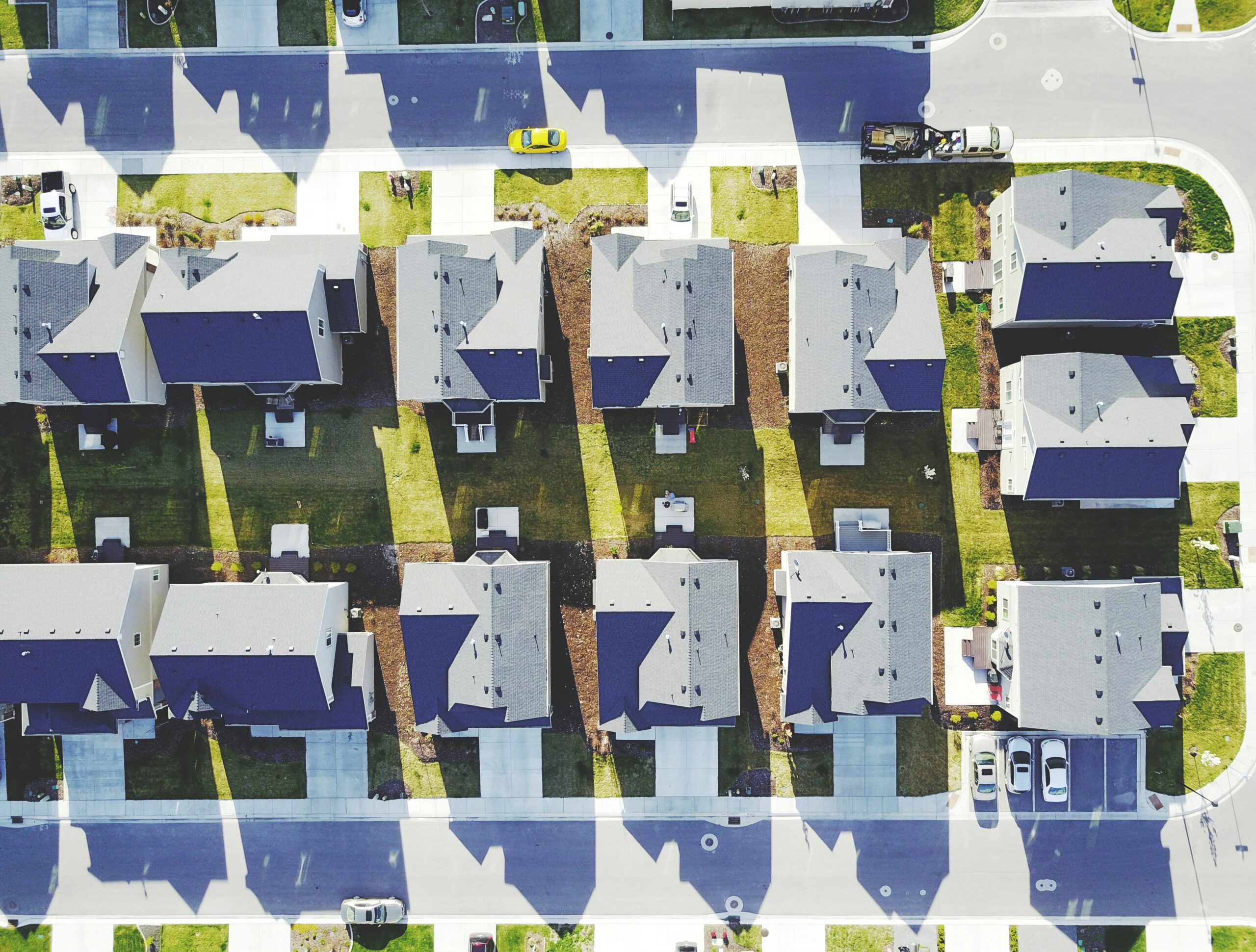 Aerial view of a suburban neighborhood showing rows of houses with driveways, lawns, and parked cars along the streets on a sunny day.