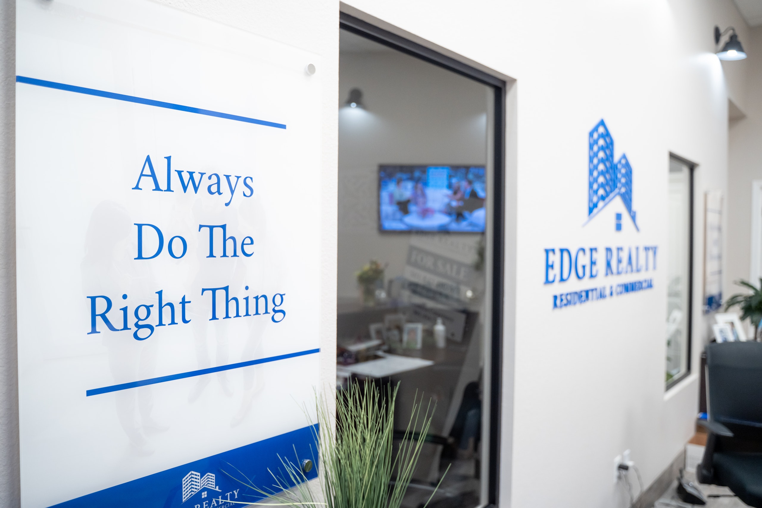 A sign on a white wall reads “Always Do The Right Thing” in blue text. Next to it is a glass office window labeled “EDGE REALTY Residential & Commercial.” Office furniture and a TV are visible inside.