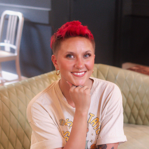 A smiling person with short, bright pink hair sits on a light-colored quilted sofa, resting their chin on their hand, wearing a beige T-shirt and earrings. A chair and dark wall are visible in the background.