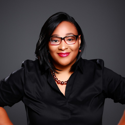 A confident person with shoulder-length black hair, wearing glasses, a black blouse, a beaded necklace, and red lipstick, smiling against a plain dark background.