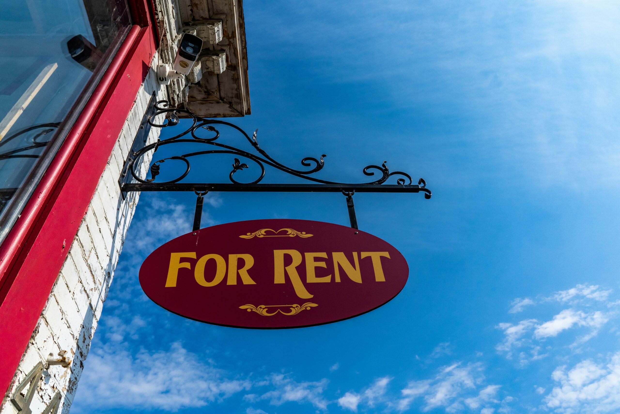 A red oval For Rent sign with yellow letters hangs outside a building against a bright blue sky. The sign, great for those considering lease vs rent options, is attached to an ornate black metal bracket above a red doorframe.
