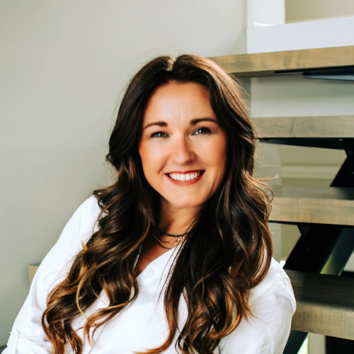 A woman with long, wavy brown hair and a white shirt smiles while sitting on wooden stairs in a modern setting.