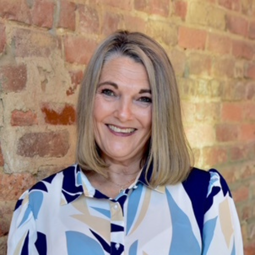 A woman with shoulder-length blonde hair smiles at the camera. She is wearing a patterned blouse with blue, beige, and white colors, standing in front of a brick wall.