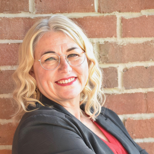 A smiling woman with wavy blonde hair, glasses, and red lipstick leans against a brick wall, wearing a dark blazer over a red top.
