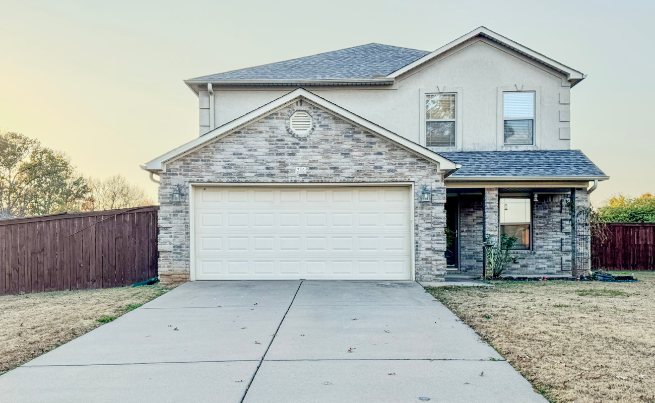 A two-story suburban house with a brick and stucco exterior, a white double garage door, and a driveway. The yard has dry grass, and a wooden fence borders the property.