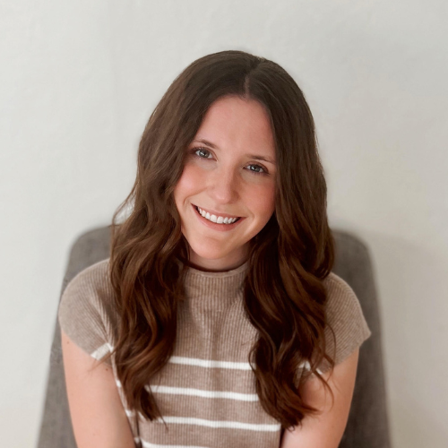 A woman with long wavy brown hair smiles at the camera. She is wearing a short-sleeve beige top with white horizontal stripes and is seated against a plain light-colored background.
