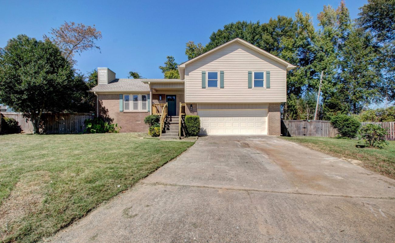 A beige two-story house with a brick lower level, a front porch, and an attached two-car garage. The house sits on a grassy lawn with a concrete driveway and is surrounded by trees and a wooden fence.