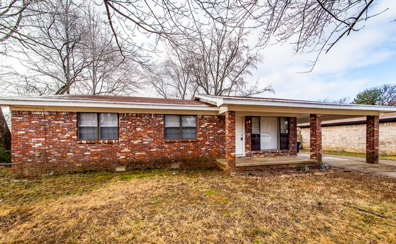 Single-story brick house with a covered front porch and attached carport. The yard has dry grass and leafless trees, indicating winter or early spring. The sky is partly cloudy.
