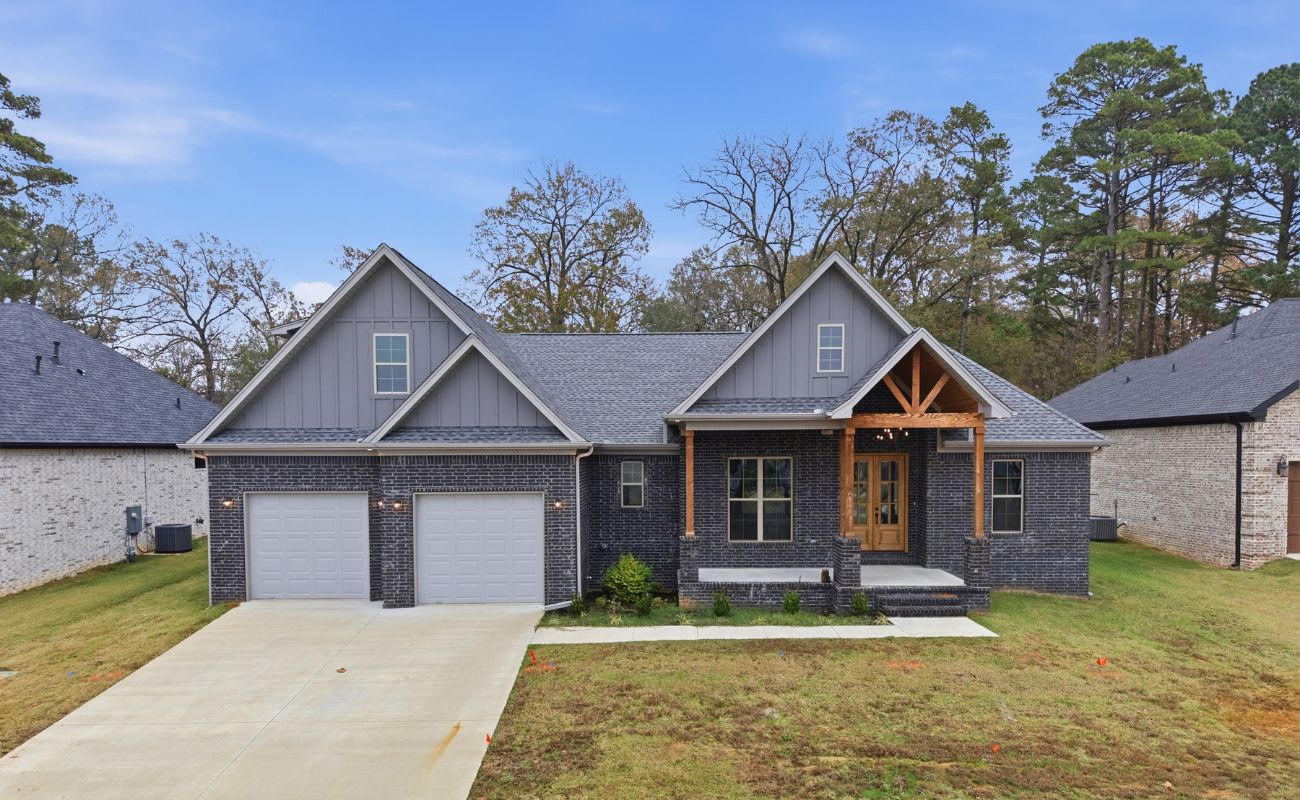 A modern, single-story house with dark brick exterior, double garage, large front porch with wooden beams, and gabled roof, set on a grassy yard with trees in the background.