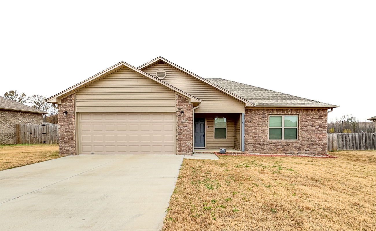 A single-story suburban house with a two-car garage, brick and siding exterior, a blue front door, and a paved driveway, surrounded by a dry grass lawn and a wooden backyard fence.