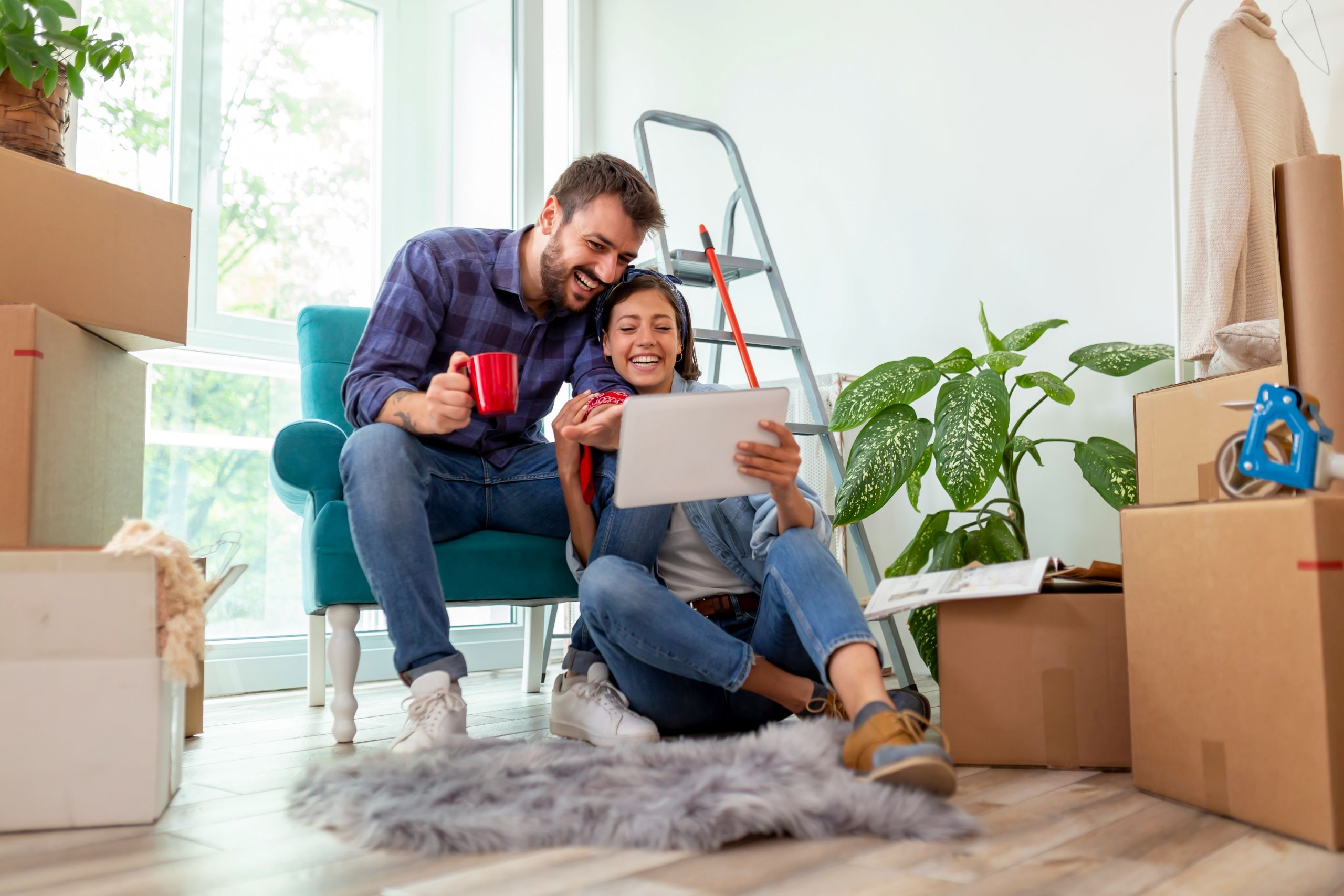 A smiling Arkansas renters couple sits among moving boxes in a bright room, looking at a tablet together. The man holds a red mug while the woman laughs, and a ladder and houseplants are visible in the background.
