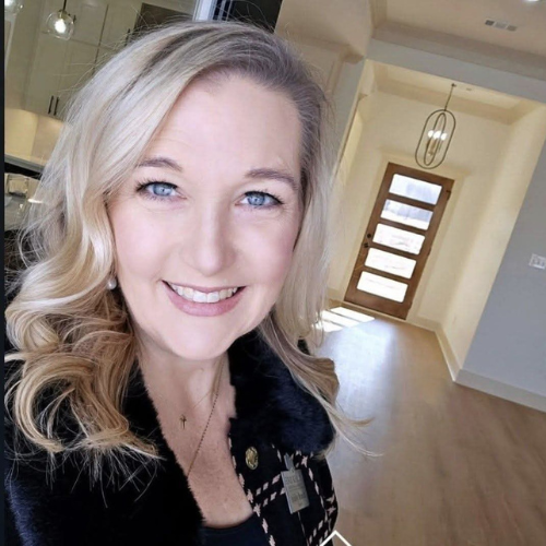 A smiling woman with wavy blonde hair stands indoors near the entrance of a modern, well-lit home with wooden floors and a glass front door. She is wearing a dark jacket with a plaid pattern and a name badge.