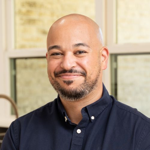 A bald man with a short beard and mustache, wearing a navy blue button-up shirt, smiles at the camera. The background shows a window with blurred light and a beige wall.