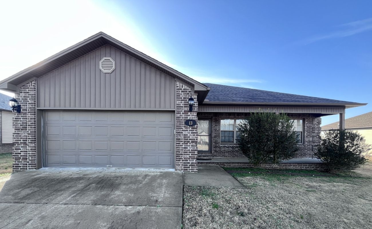 A single-story house with gray brick and siding, a two-car garage, two front bushes, and a concrete driveway, set under a clear blue sky.