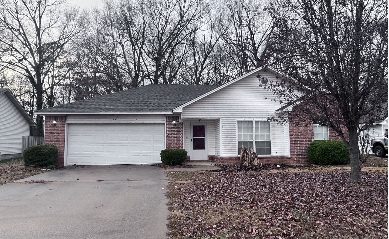 A single-story brick and white-sided house with a two-car garage, a leafless tree in the front yard, and brown leaves scattered on the driveway and lawn. Overcast sky and bare trees in the background.