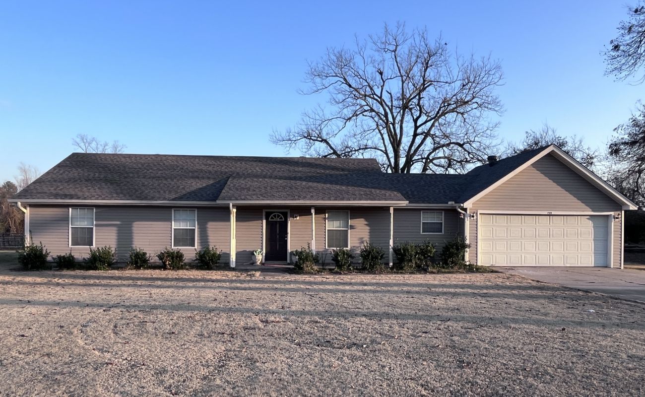 Single-story beige house with dark roof, small covered porch, white-framed windows, attached two-car garage, and a row of shrubs in front. Leafless tree stands behind the house under a clear blue sky.