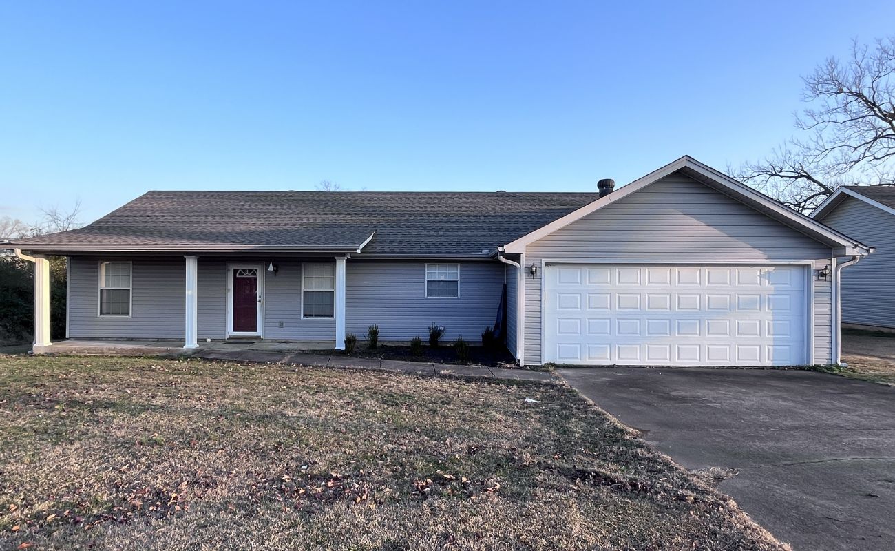 A single-story house with gray siding, a red front door, white trim, and a double garage. The front yard has patches of grass and a concrete driveway. The sky is clear and blue.