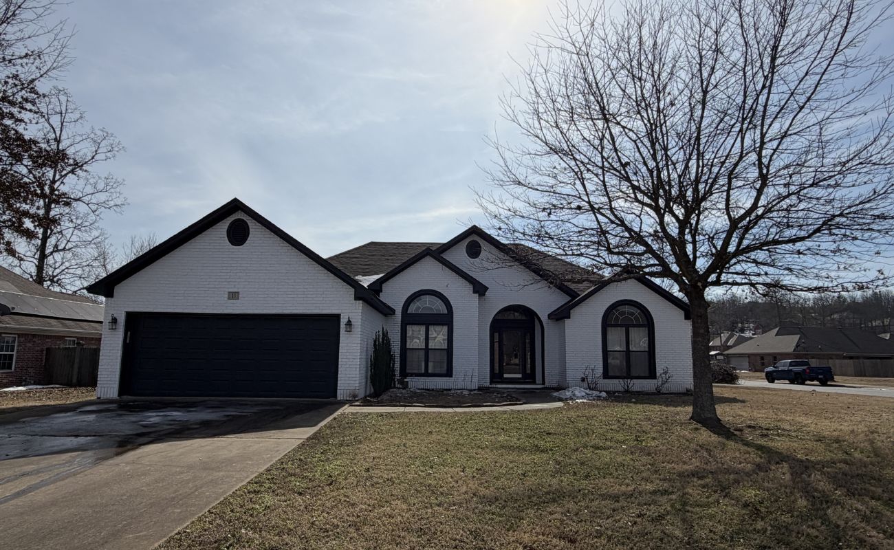 A white brick single-story house with a dark garage door, arched windows, and a leafless tree in the front yard, set on a corner lot with a clear sky overhead.