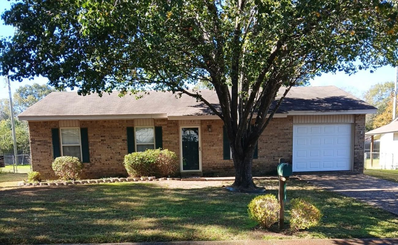 A one-story brick house with green shutters, a single-car garage, a large tree in the front yard, and a green mailbox near the driveway. The lawn is partly shaded by the tree.