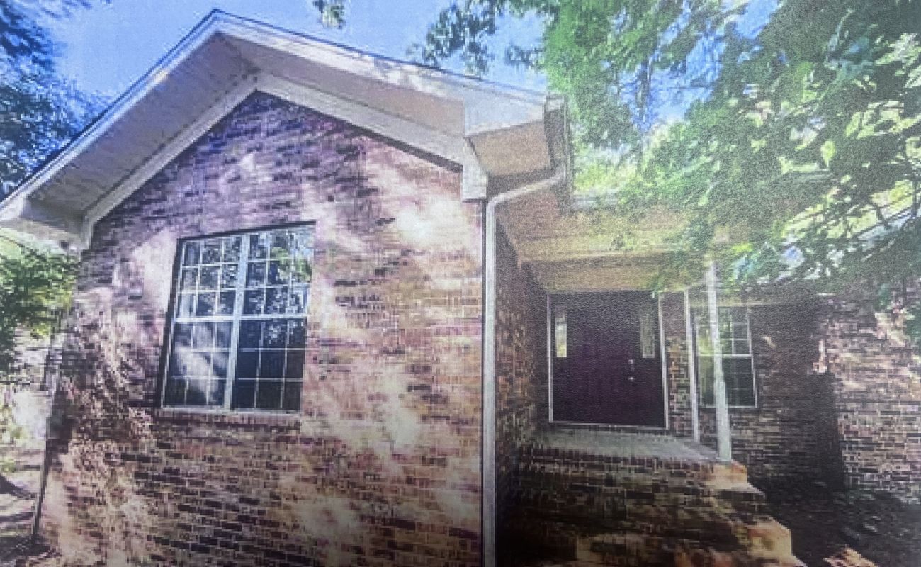 A brick house with a large front window, a covered entryway, and steps leading to a dark-colored front door, surrounded by leafy trees casting shadows on the exterior.
