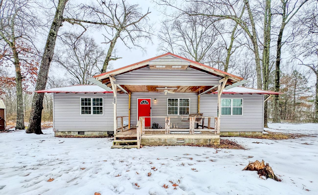A small house with gray siding, a red roof, and a bright red front door sits in a snowy yard surrounded by leafless trees. A covered porch with rocking chairs is at the front of the house.