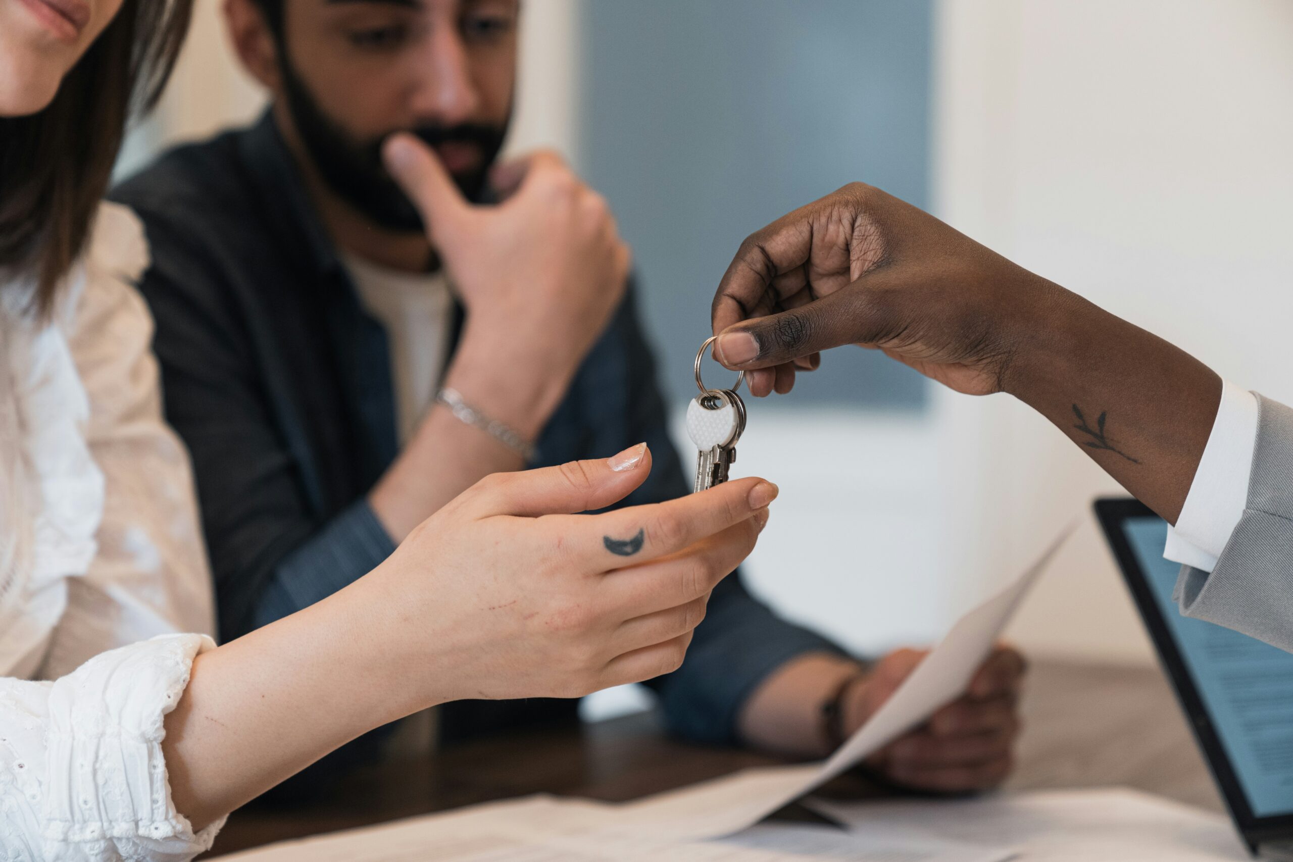 A person hands over a set of keys to another, while a third person—likely one of the property investors—watches thoughtfully. Documents and a laptop are on the table in front of them.