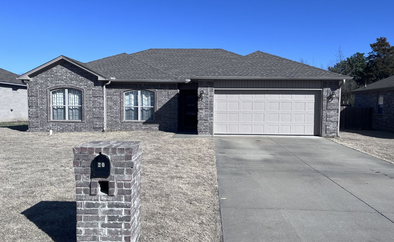Single-story brick house with a double garage, arched windows, and a mailbox labeled 20 in front. The yard has dry grass and a concrete driveway, with a clear blue sky overhead.
