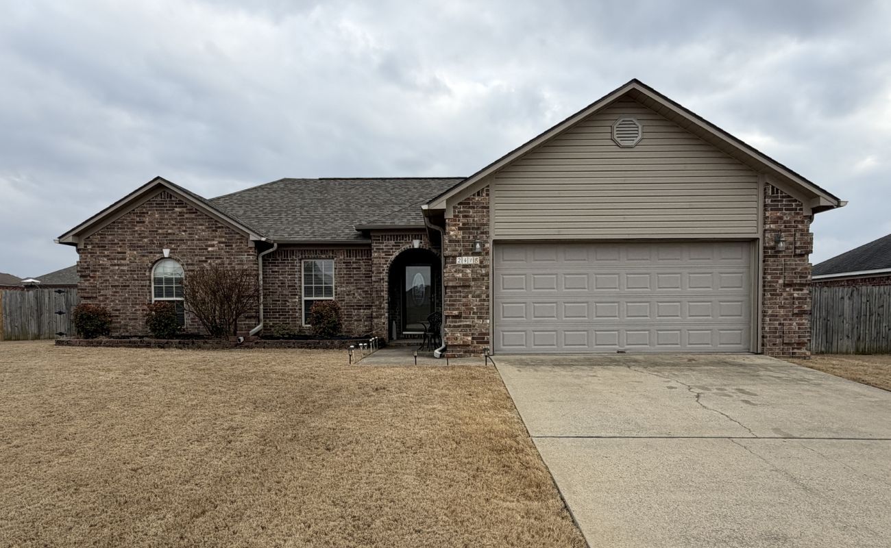 A single-story brick house with a two-car garage, a gray shingle roof, arched front entryway, and a large, dry grass lawn under an overcast sky.