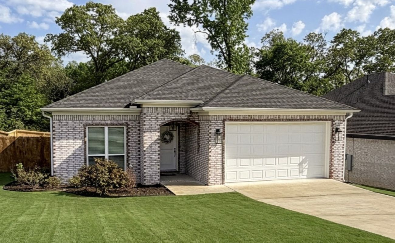 Single-story brick house with a dark gray roof, white garage door, neat landscaping, and a concrete driveway. Green lawn and trees surround the home under a partly cloudy sky.