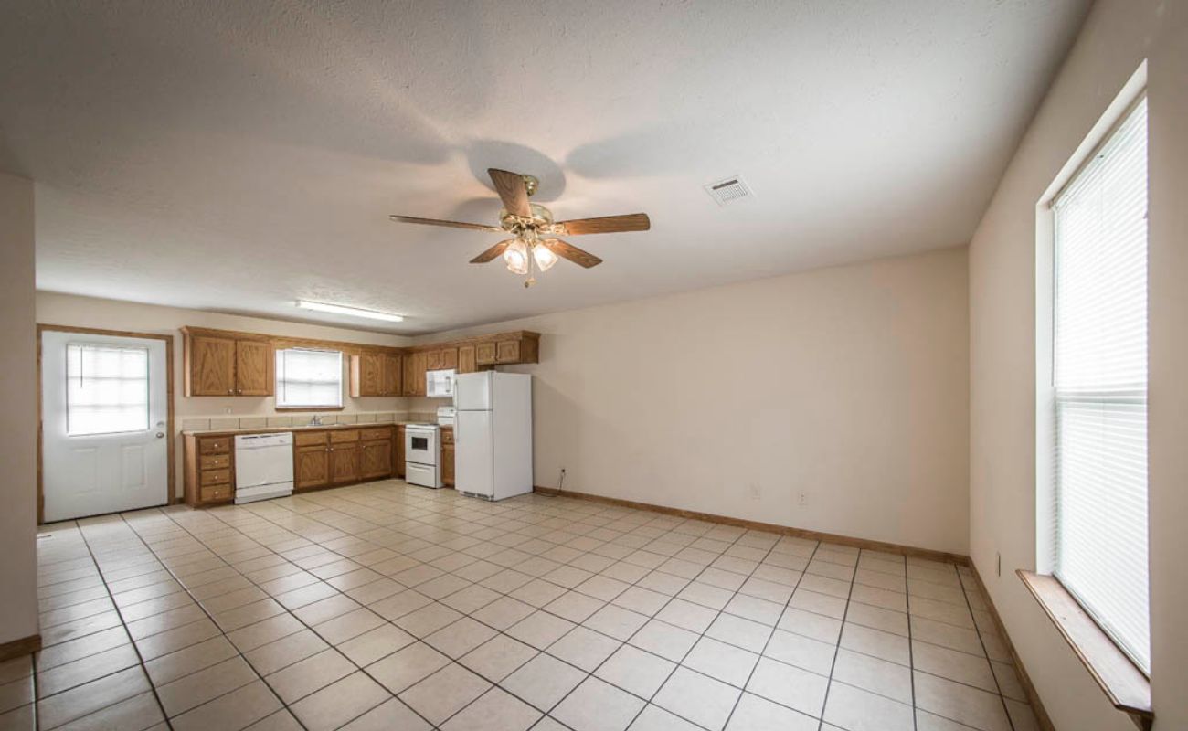 A spacious, unfurnished kitchen and living area with tiled floors, wooden cabinets, white appliances, a ceiling fan with lights, and large windows allowing natural light into the room.