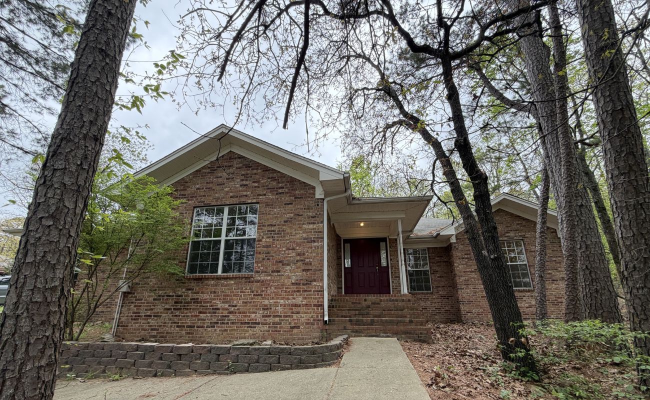 A single-story brick house with a red front door, large front window, and a small covered entryway, surrounded by tall trees and a concrete walkway leading to the entrance.