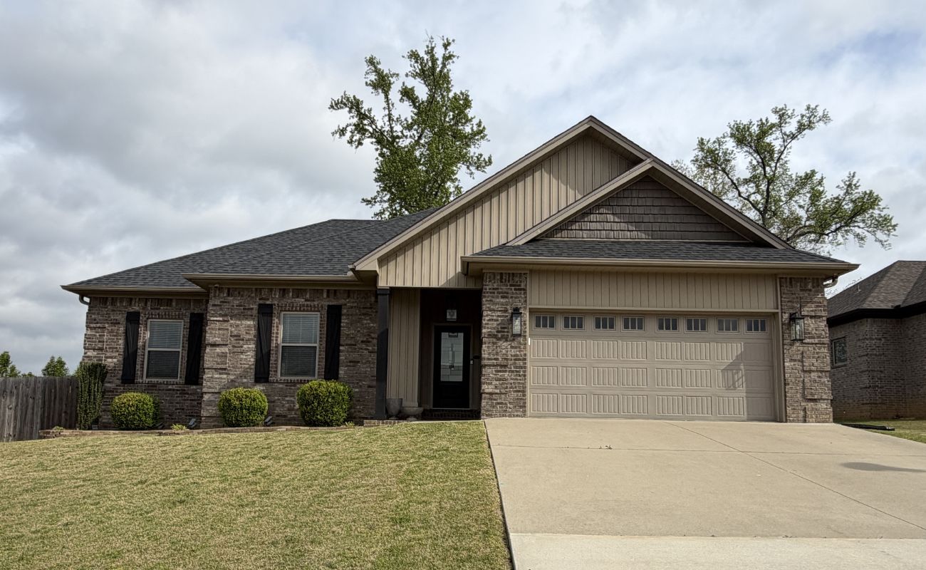 A single-story suburban house with a brick and siding exterior, black shutters, a two-car garage, and a sloped driveway, set on a grassy lawn with neatly trimmed bushes.