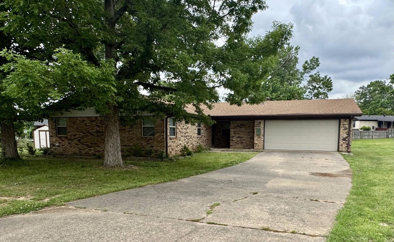 A single-story brick house with a brown roof and attached two-car garage. A large tree and green lawn are in the front yard, and a wide driveway leads to the garage. The sky is cloudy.