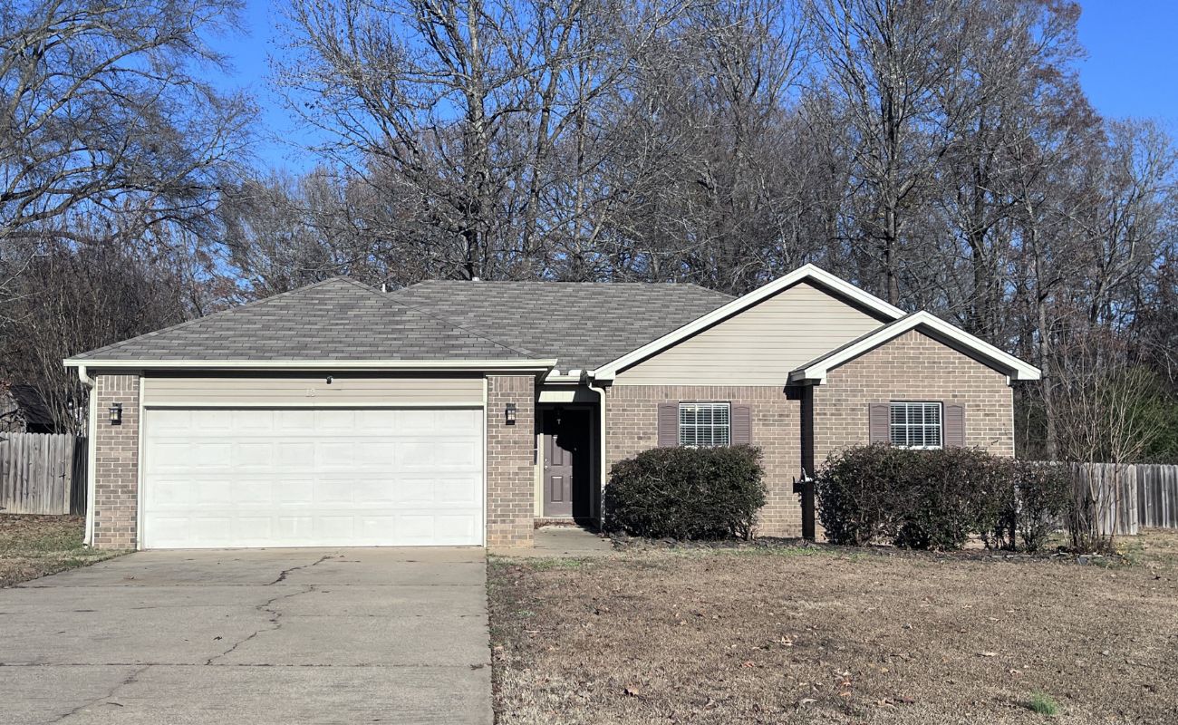 A single-story brick house with a two-car garage, small front bushes, and a bare lawn. Leafless trees and a wooden fence are in the background under a clear blue sky.