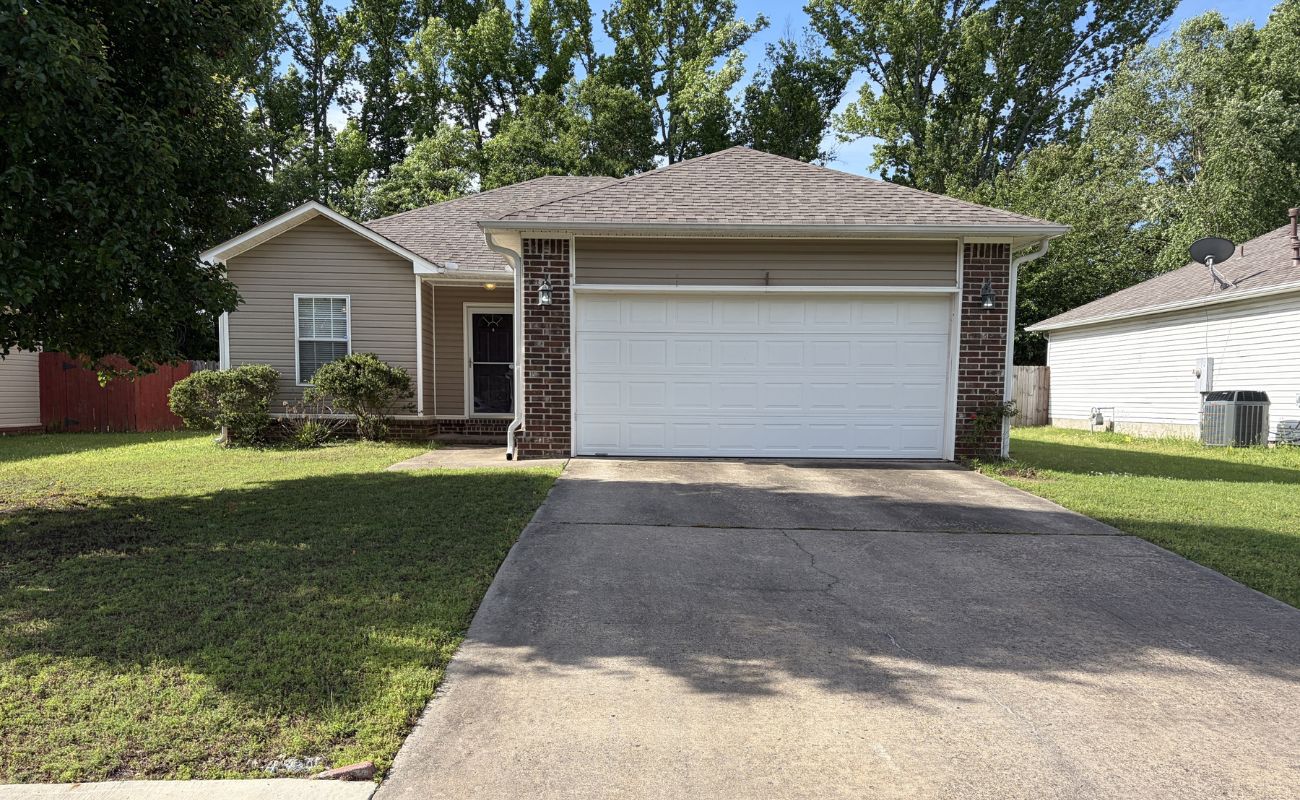 A single-story house with tan siding and brick accents, a white two-car garage, and a concrete driveway. The lawn is green with some bushes near the front door, and trees are in the background.