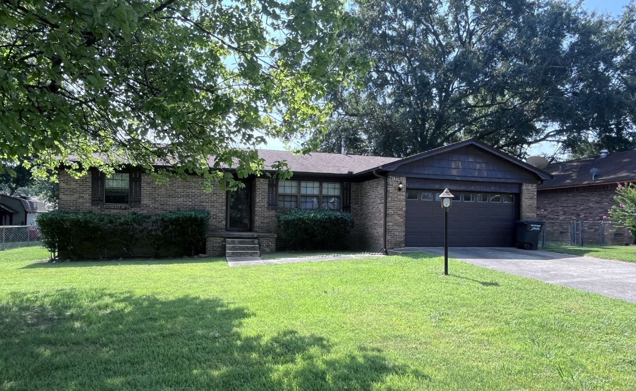 Single-story brick house with dark trim, shrubbery in front, a two-car garage, and a concrete driveway. A lamp post stands in the front yard, with trees and green grass surrounding the home.