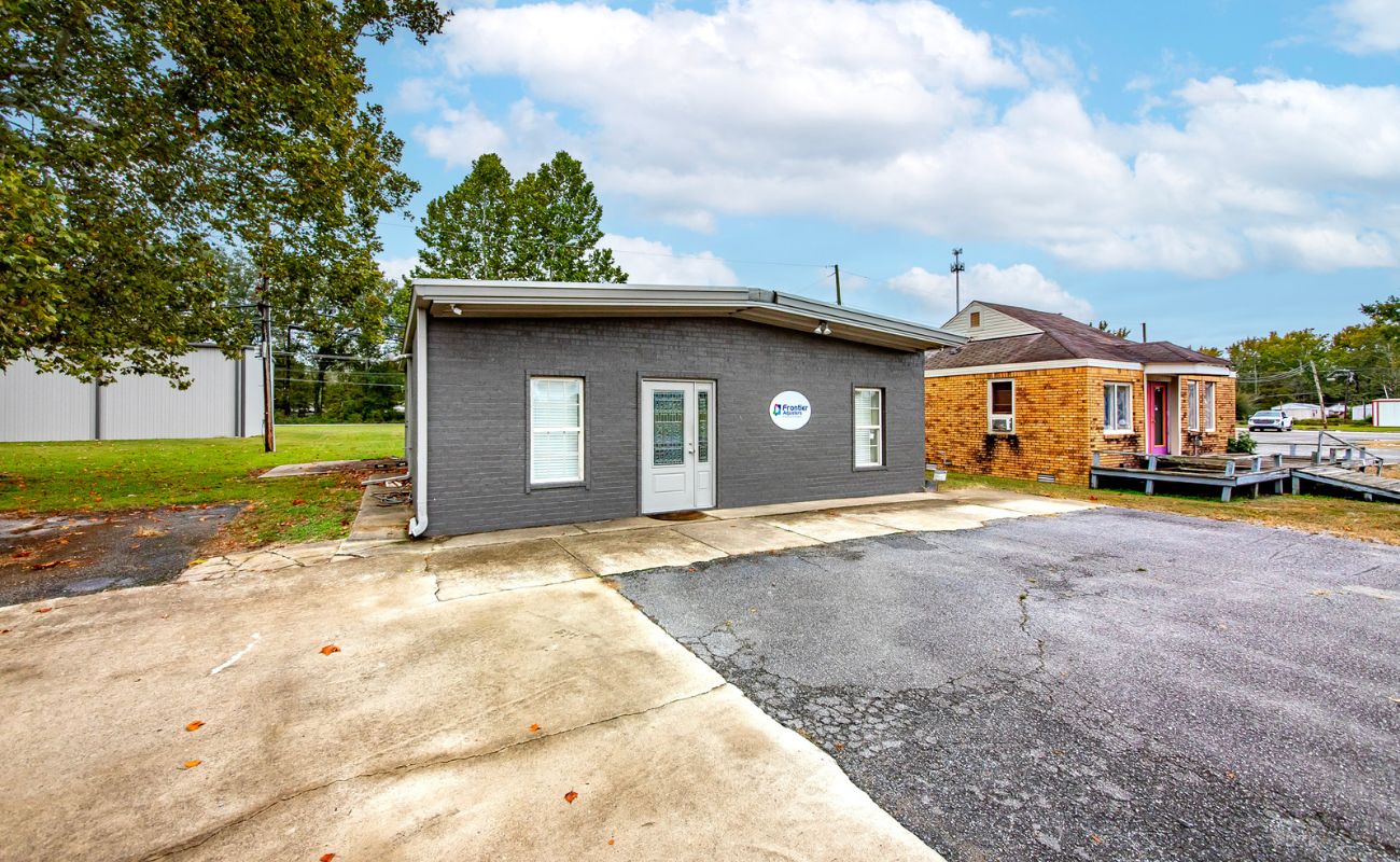 A small, gray brick office building with two front doors and a circular sign, next to a tan house, surrounded by trees and a large, cracked concrete parking area under a partly cloudy sky.